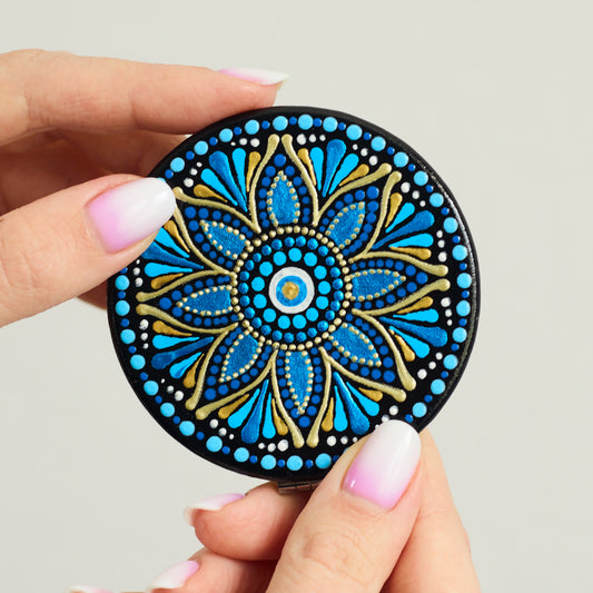 Round compact mirror with blue  mandala design held by a hand on a white background