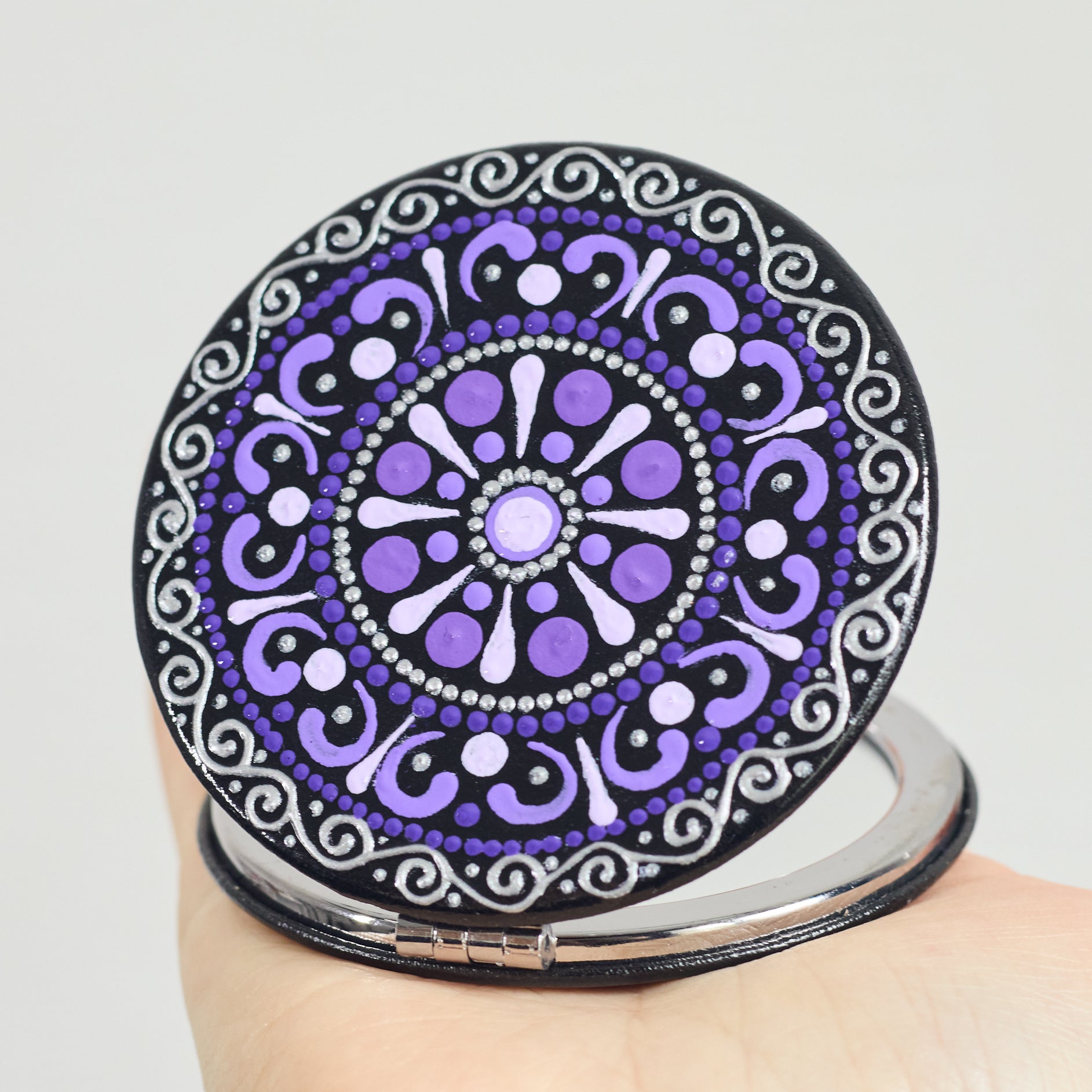 Round compact mirror with purple  mandala design held by a hand on a white background
