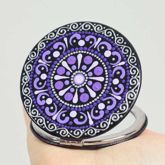 Round compact mirror with purple  mandala design held by a hand on a white background