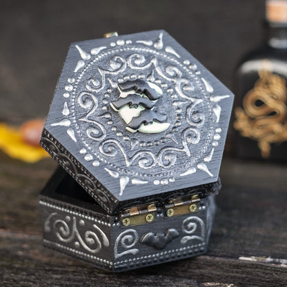 A hexagonal black wooden box with bats on the moon polymer clay figurine attached to the top of the box. Hand-painted designs in silver. Poison bottles on the wooden background.