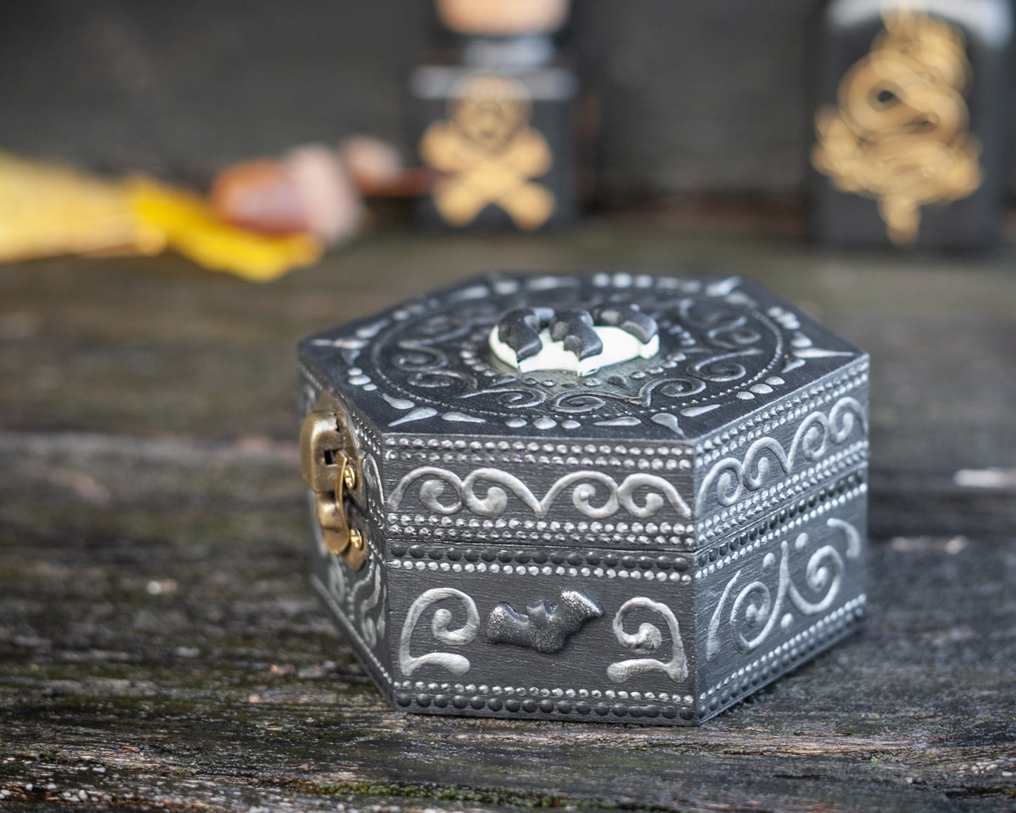 A hexagonal black wooden box with bats on the moon polymer clay figurine attached to the top of the box. Hand-painted designs in silver. Poison bottles on the wooden background. View from the side.