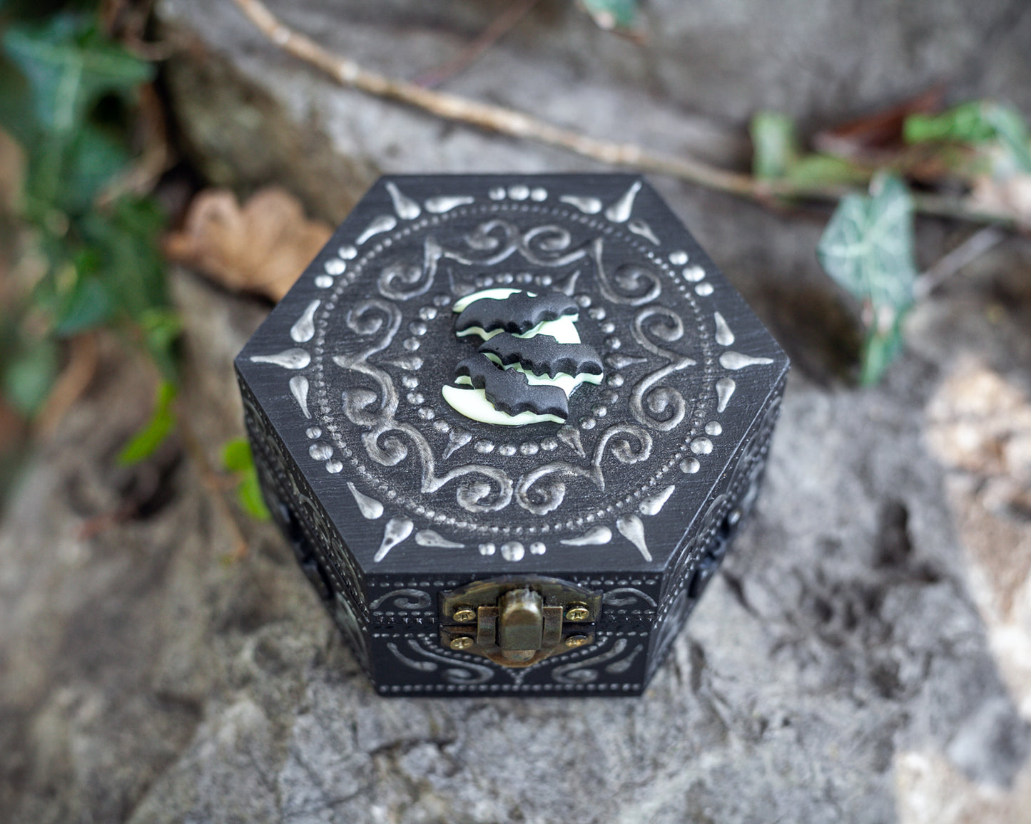 A hexagonal black wooden box with bats on the moon polymer clay figurine attached to the top of the box. Hand-painted designs in silver. The box sits on a rock.