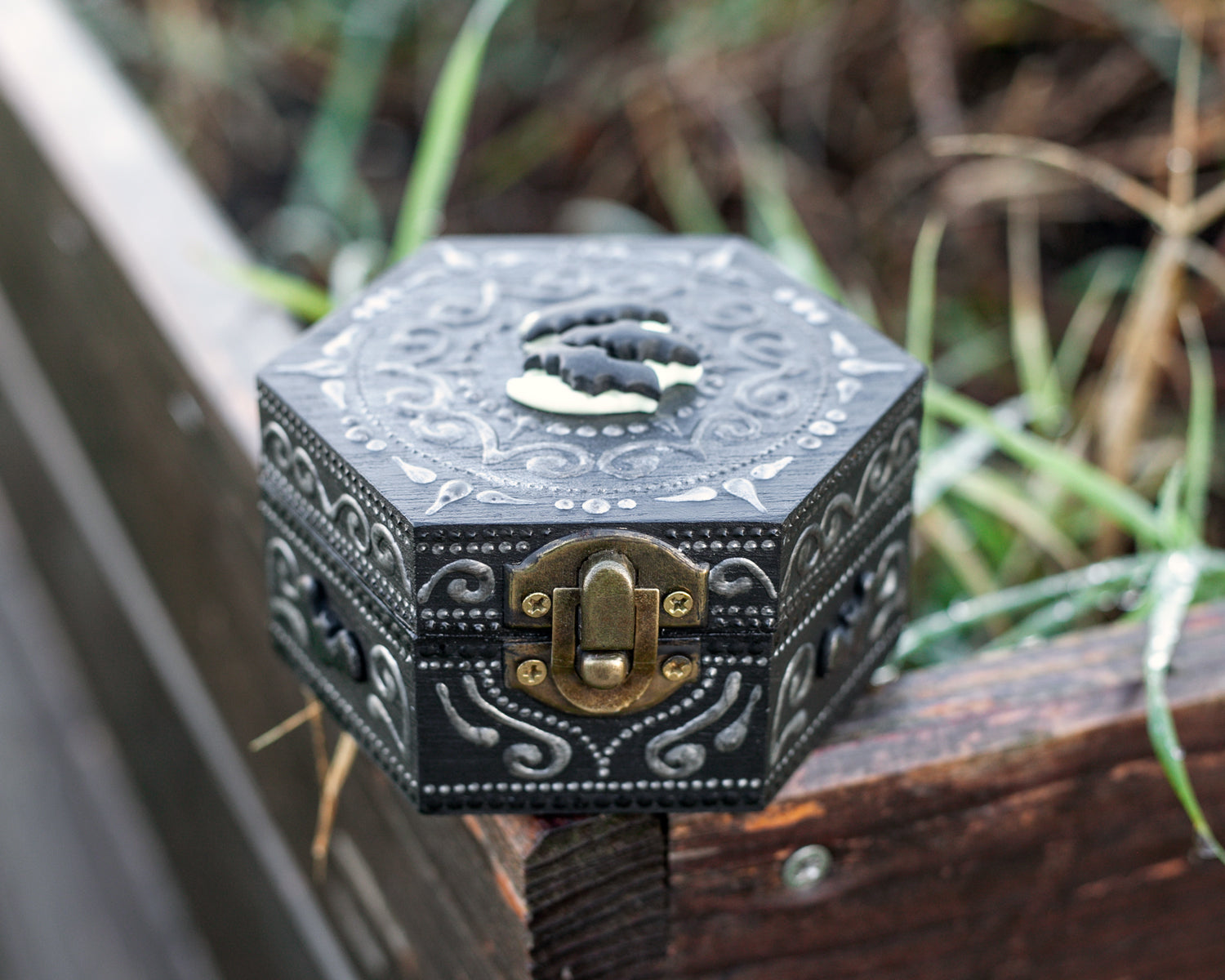 A hexagonal black wooden box with bats on the moon polymer clay figurine attached to the top of the box. The box sits on a rock. Close-up front view to show the metal closure.