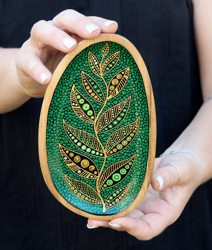 Hands holding small green wooden plate with a golden leaf design, black background.