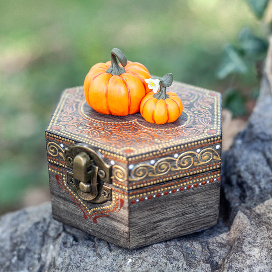 A hexagonal wooden box with hand-painted designs in gold and red sits on a mossy rock. Two miniature pumpkins, sculpted from polymer clay with remarkable detail, are attached to the top of the box. The background is a soft focus of green foliage.
