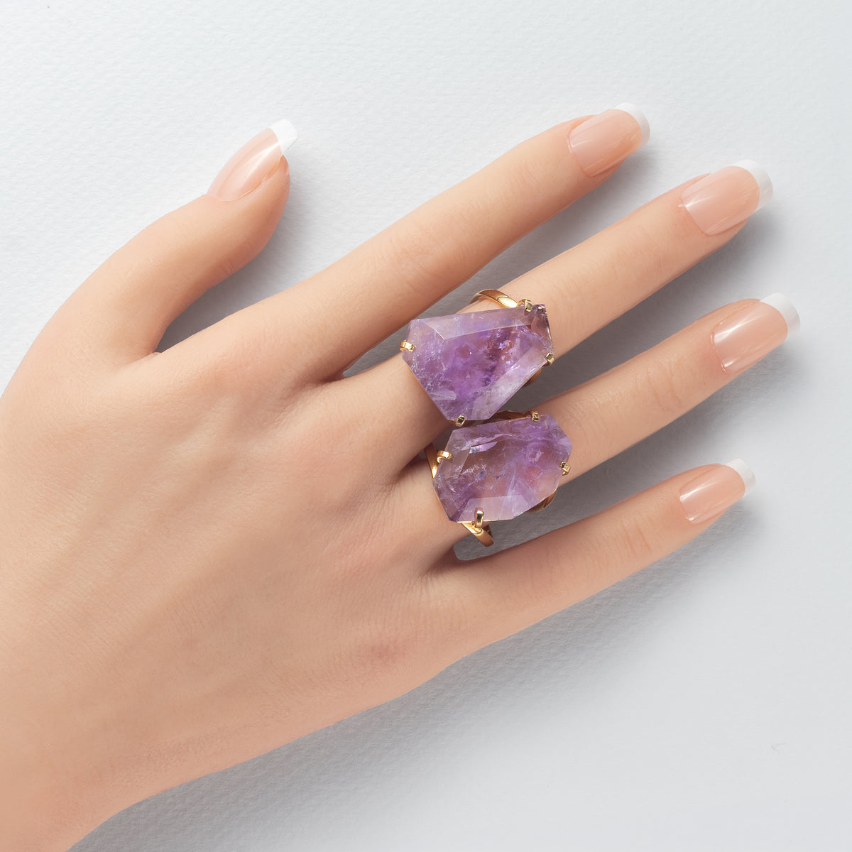 A close-up of a hand wearing two gold rings, each featuring a large, rough-cut purple amethyst gemstone, set against a light gray background.