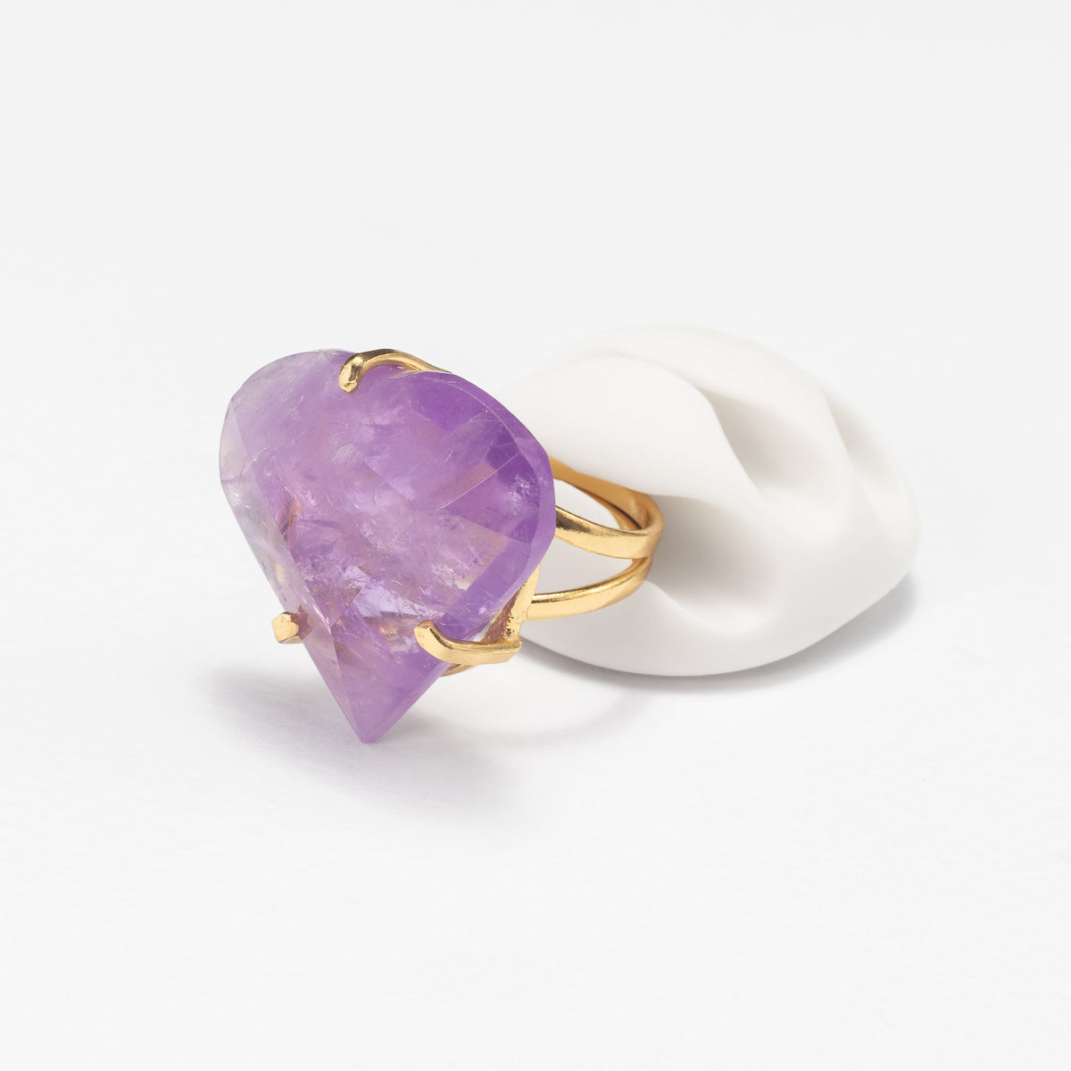 A gold ring featuring a large, faceted purple gemstone, displayed on a white sculptural ear stand against a clean white background.