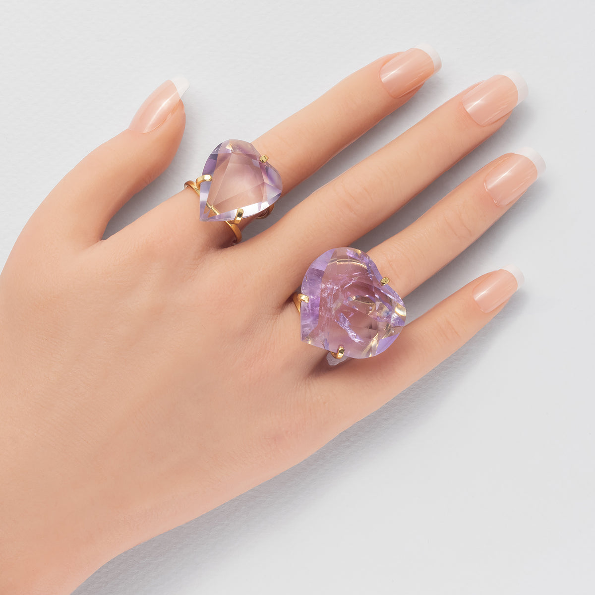 A close-up of a hand wearing two heart-shaped purple amethyst gemstone rings with gold settings against a light gray background.