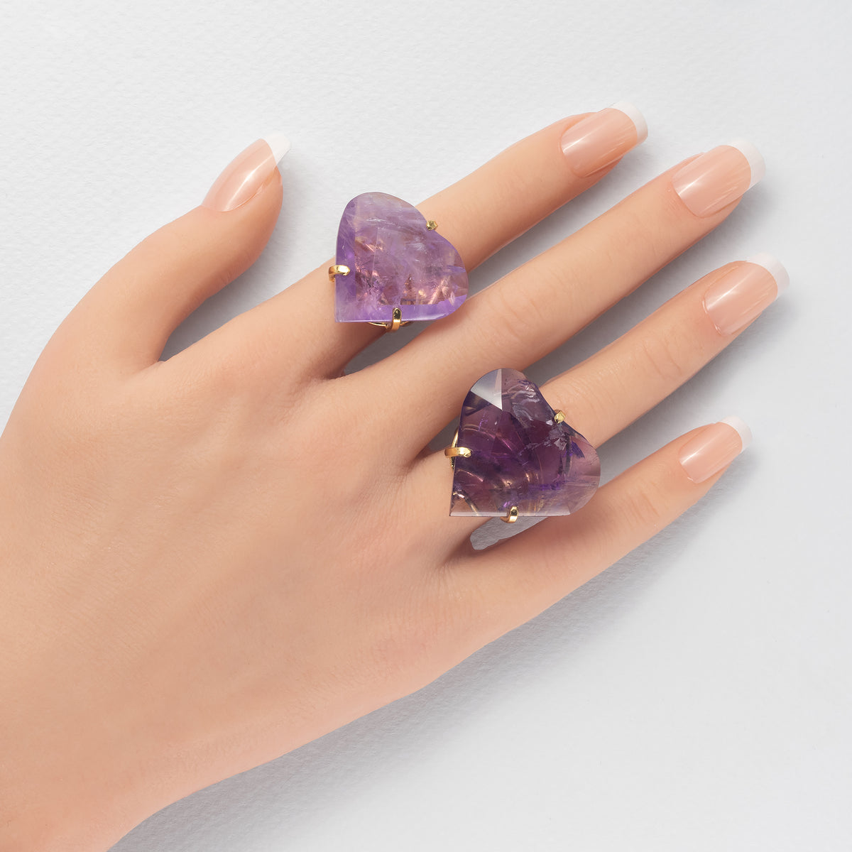 A hand close-up wearing two chunky rings featuring raw, faceted purple amethyst crystals in gold settings, set against a clean, light gray background.