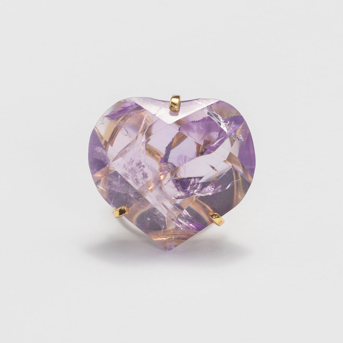 A heart-shaped faceted purple gemstone in a gold rings setting, photographed against a clean light gray background.