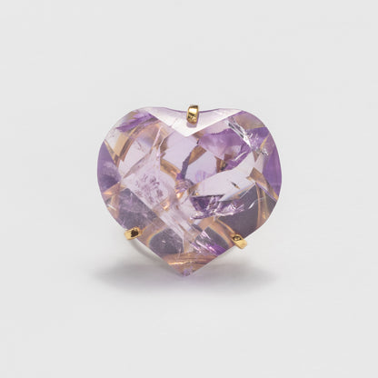 A heart-shaped faceted purple gemstone in a gold rings setting, photographed against a clean light gray background.
