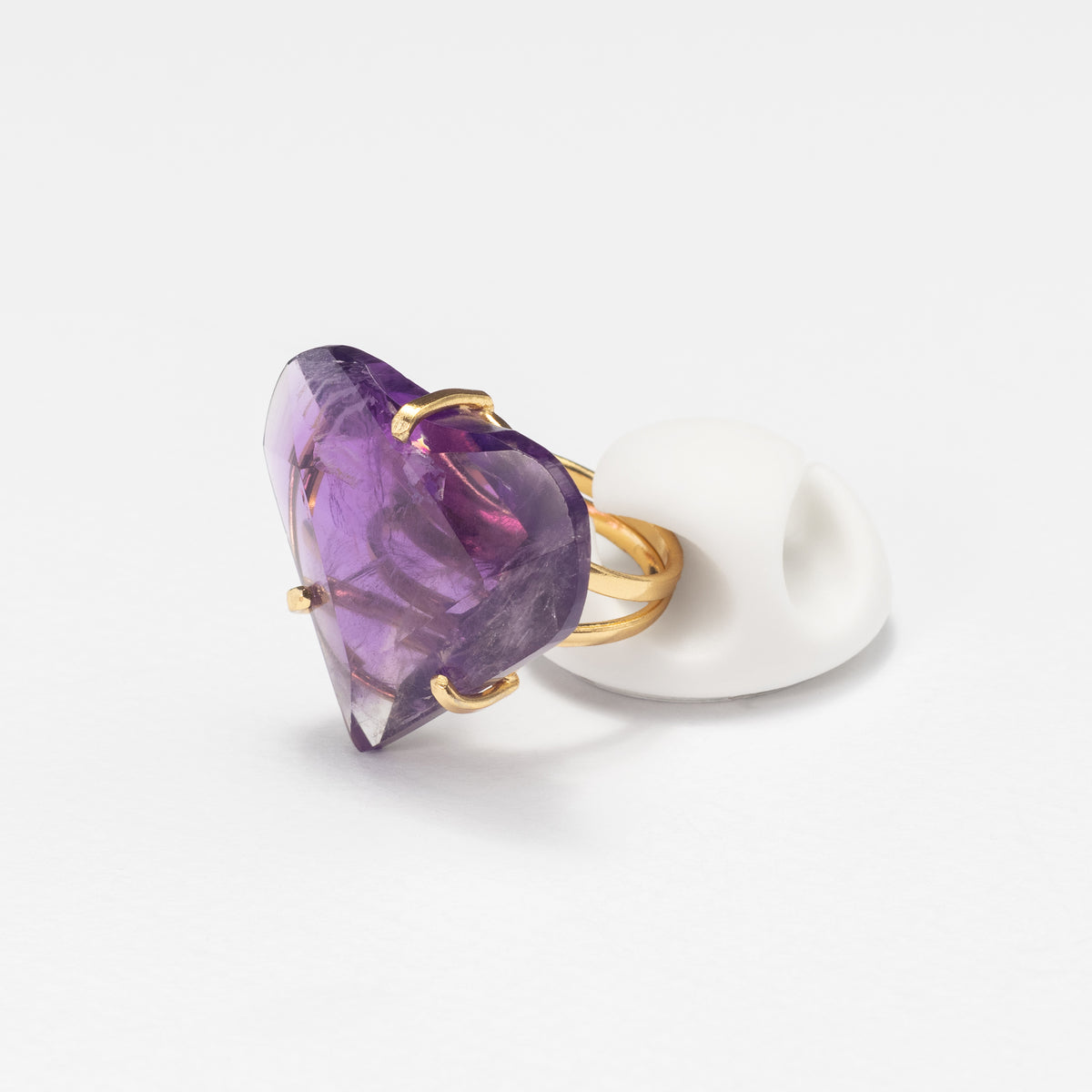 A heart-shaped purple amethyst ring with gold metal prongs and band, displayed on a white ring stand against a clean white background.