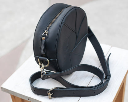 Black leather round bag on a white surface with a light gray background. Close-up view from the side to show a zipper closure.
