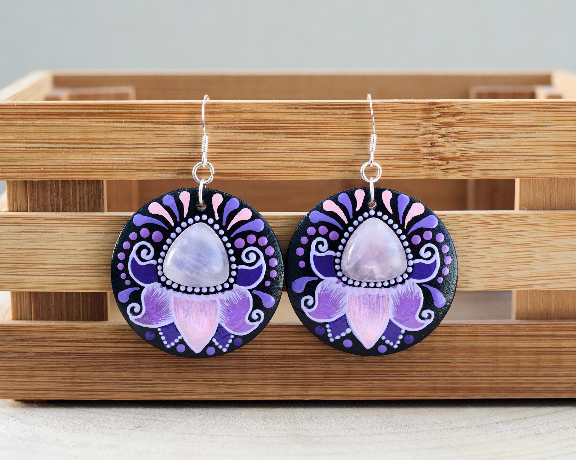 Wooden earrings with purple and pink floral design and rose quartz cabochon on a wooden stand