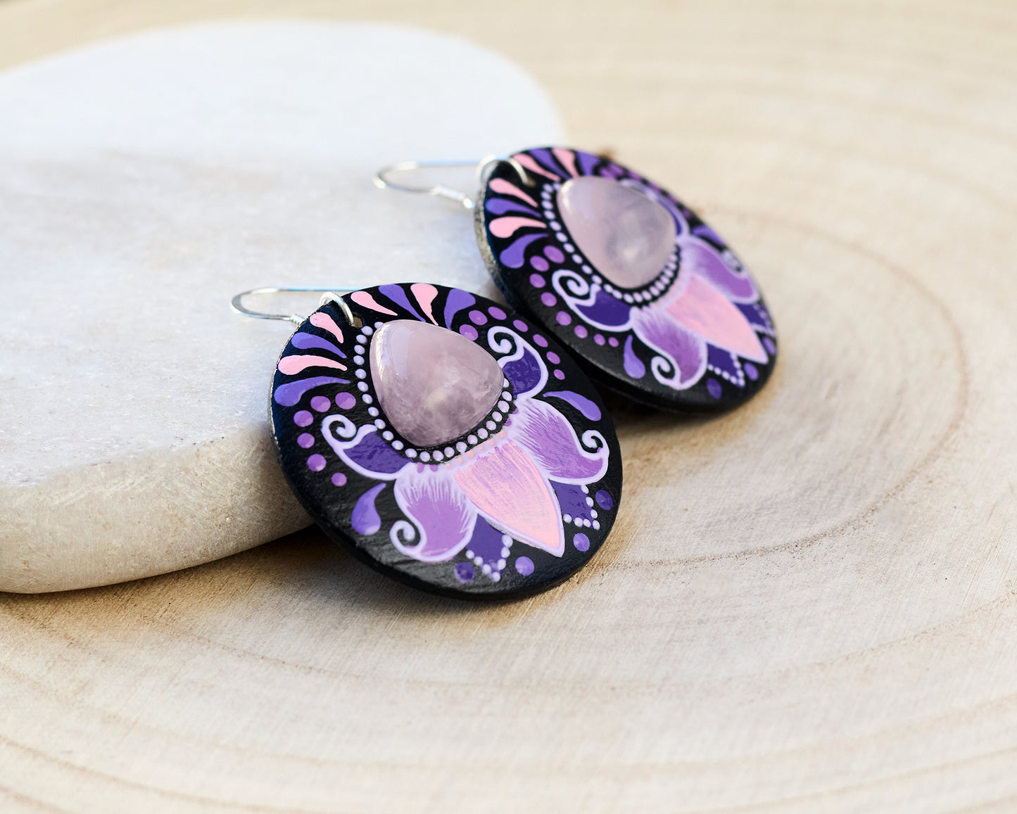 Pair of wooden earrings with purple and pink floral design and rose quartz cabochon on a light wooden surface.