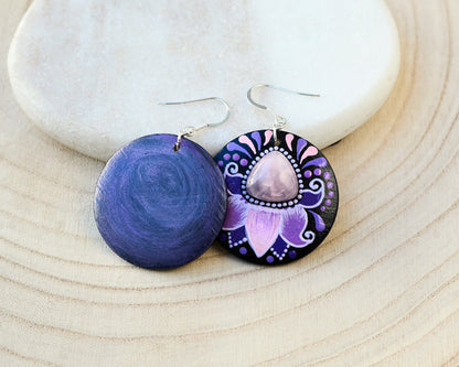 Pair of wooden earrings with purple and pink floral design and rose quartz cabochon on a light wooden surface.