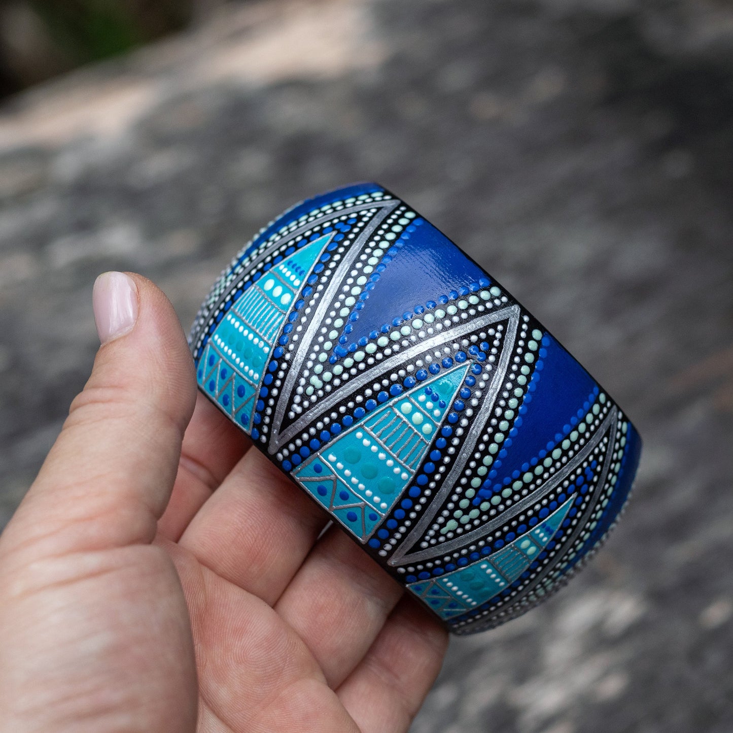 Hand holding a blue wide patterned bracelet against a blurred natural background