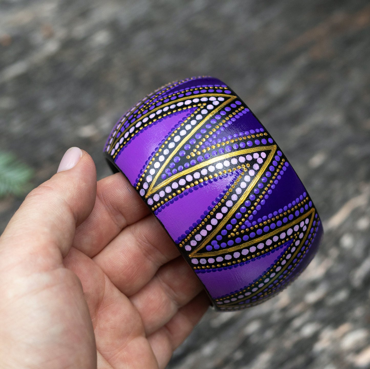 Hand holding a purple wide patterned bracelet against a blurred natural background
