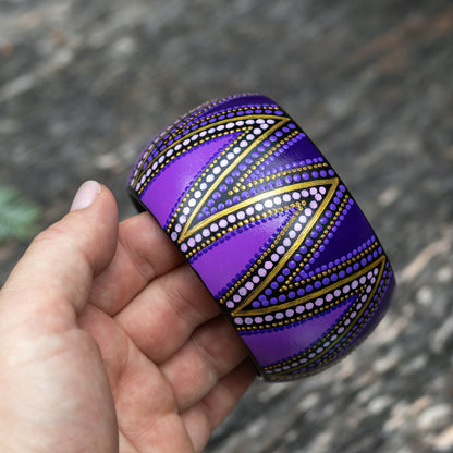 Hand holding a purple wide patterned bracelet against a blurred natural background