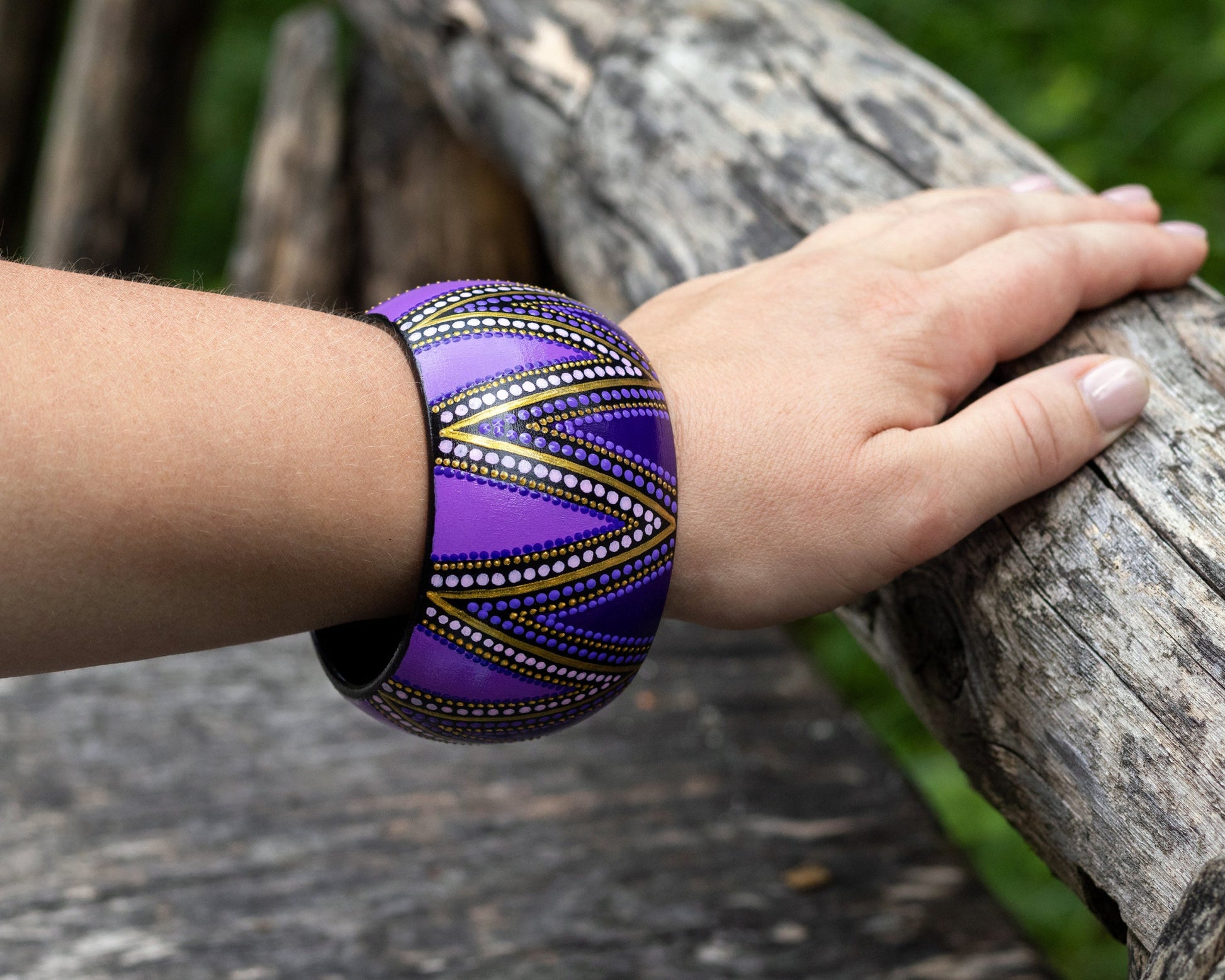 Hand wearing a purple wide bracelet with gold tones, holding onto a wooden log.