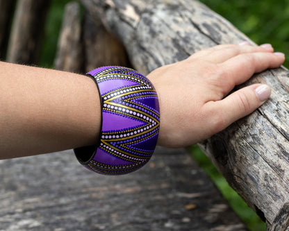 Hand wearing a purple wide bracelet with gold tones, holding onto a wooden log.