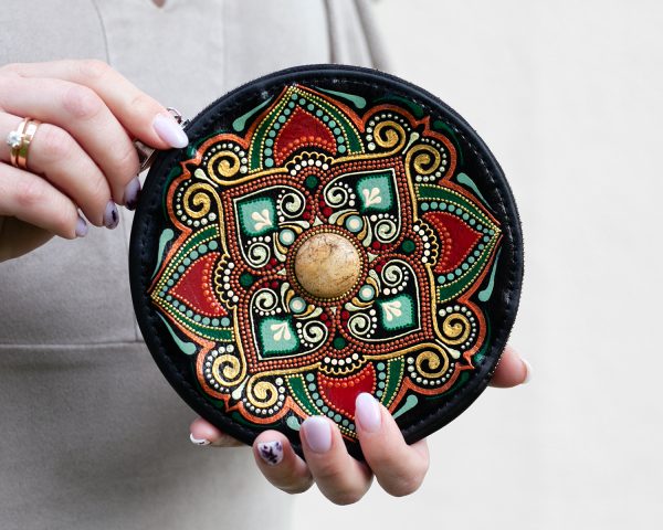 Woman holding in her hands round leather wallet with ethnic painting in earth colors and natural jasper inlay in the middle. Close-up view